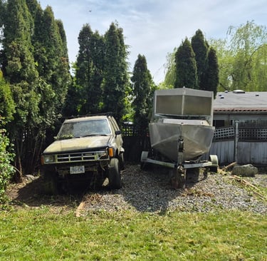 an aluminum boat on a trailer parked beside an older Toyota SUV. Both on a gravel pad in front of an older fence