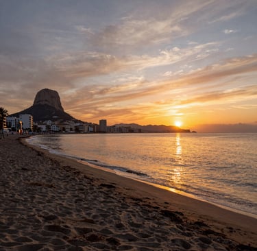 Golden sunrise over a sandy Mediterranean beach with the Peñón de Ifach rock formation in Calpe, Spain.