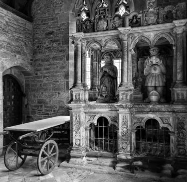 Black and white interior of a stone church featuring ornate gothic tombs, statues, and a wooden cart.