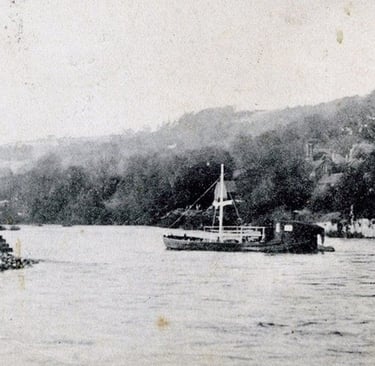 Vintage black and white photograph of a small sailing boat on a river near a wooded shoreline.