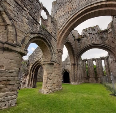 Ancient stone ruins of a medieval abbey with gothic arches and grassy interior under a cloudy sky.
