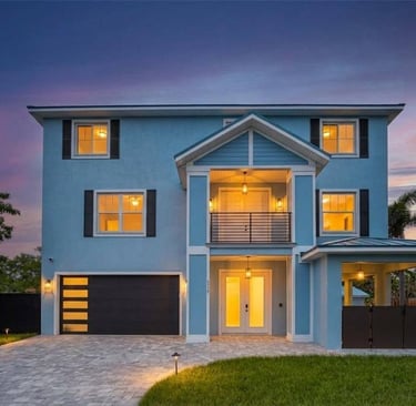 A modern light blue two-story home with a paved driveway and warm exterior lighting at sunset.