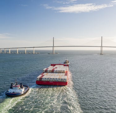 Aerial view of a red cargo ship and tugboat passing under the Sunshine Skyway Bridge in Florida.