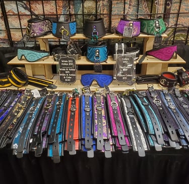 A display of colorful leather BDSM collars, wrist cuffs, and lace eye masks at a craft fair.