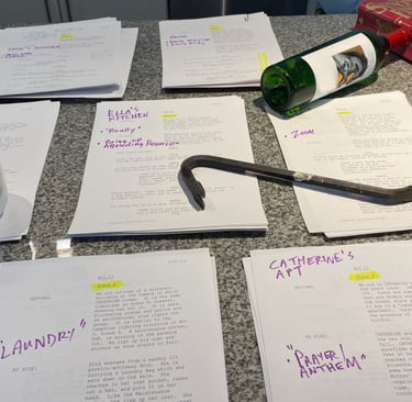 Multiple piles of play script on a table, with a coffe cup, empty wine bottle & crowbar on top