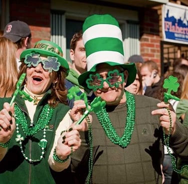 People celebrating St. Patrick's Day wearing green hats, shamrock glasses, and beads at a festive parade.