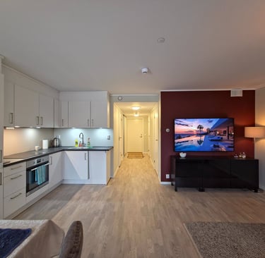 Modern open plan apartment featuring a white kitchen and living area with a wall-mounted TV on a red accent wall.