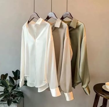 Elegant long-sleeve silk button-down blouses in white, beige, and olive green hanging on wooden hangers.