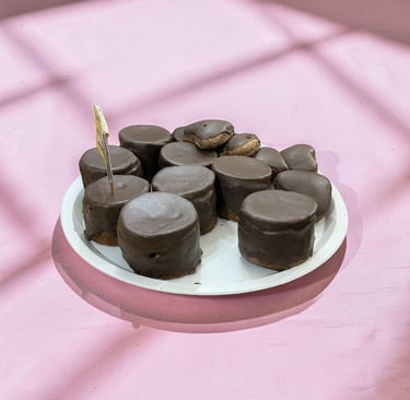Un plato blanco lleno de malvaviscos gourmet cubiertos de chocolate sobre una superficie rosa con so