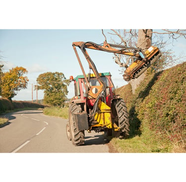 A tractor with a mechanical flail arm attachment trims a tall roadside hedge and trees.
