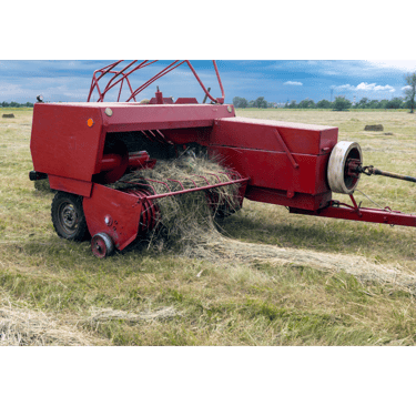 Red square hay baler harvesting dry grass in a rural field under a blue sky.