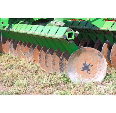 A green agricultural disc harrow with steel blades tilling dry soil in a farm field.