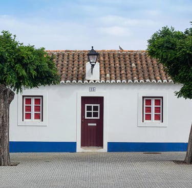 Traditional white Portuguese house with blue trim, red windows, and symmetrical green trees.