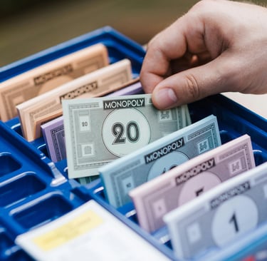 A hand selects a bill from a blue Monopoly game bank tray filled with colorful play money.