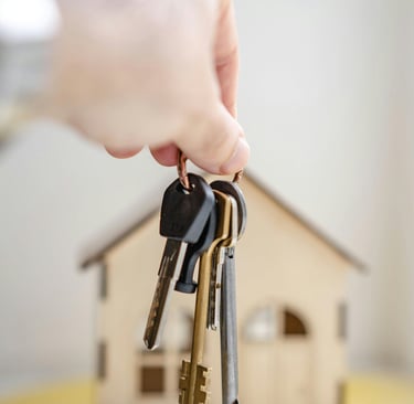 A hand holding house keys in front of a small model home, representing real estate and home ownership.