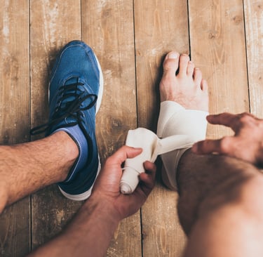 A man applying a white compression bandage to his sprained ankle on a wooden floor.