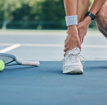 Tennis player holding an injured ankle on a blue court with a racket and ball nearby.