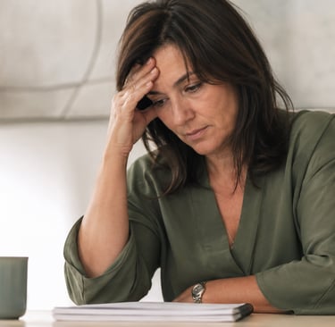 A stressed middle-aged woman sitting at a desk and holding her head while looking at documents.