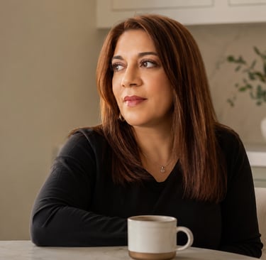 A thoughtful woman with brown hair sits in a bright kitchen with a coffee mug.