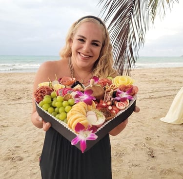 Una mujer sonriente en una playa tropical sosteniendo una tabla de charcutería en forma de corazón.