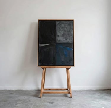 Una fotografía de estilo editorial de un estudio de artista contemporáneo en Latinoamérica. Un espacio de techos altos con paredes blancas inmaculadas y suelo de hormigón gris claro. Un caballete de madera minimalista sostiene una obra abstracta en tonos negro carbón y gris azulado bajo una luz natural suave.
