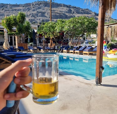 Hand holding a cold beer mug by a sunny resort swimming pool with mountain views.