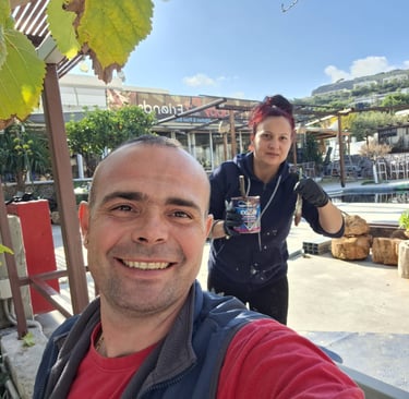 A smiling man and woman performing outdoor renovations near a swimming pool with a can of wood stain.