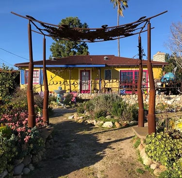 Yellow Santa Barbara home with pink trim and blue shutters, viewed through a rustic metal garden arbor.