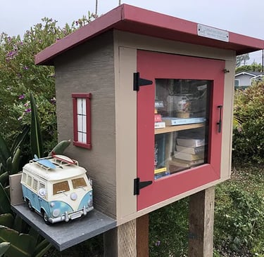 A red and tan Little Free Library book sharing box with a miniature blue VW surf van on a side shelf.