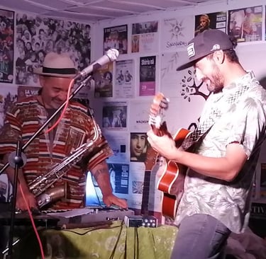 Dos músicos interpretan música en vivo con un saxofón y una guitarra acústica frente a una pared cub