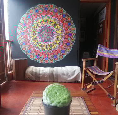 Sala con planta y mandala pintado en la pared, hay una silla mecedora y otros muebles rústicos con luz cálida