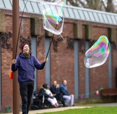 A man creates large iridescent soap bubbles outdoors using two long poles on a grassy field.