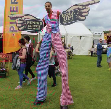 A circus stilt walker in red, white, and blue striped pants holds Pipers Circus Workshop signs at an outdoor festival.