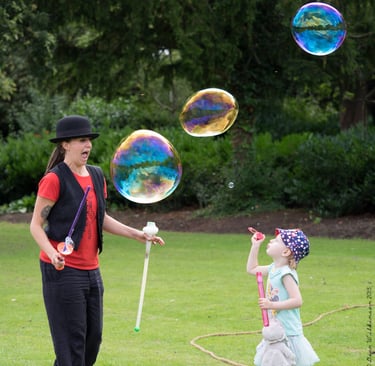 A woman and a young girl blowing large, colorful bubbles in a lush green park.