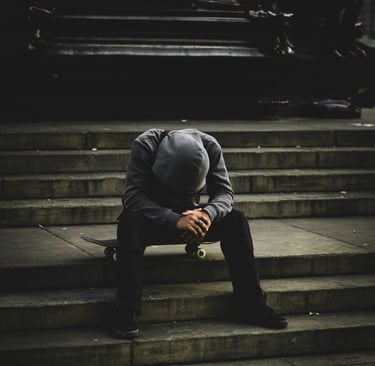 A lonely skateboarder in a grey hoodie sits on his board on city concrete stairs in a moody urban setting.