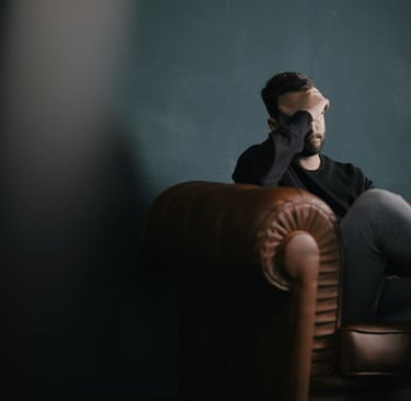 A stressed man sitting on a brown leather armchair holding his head in frustration.