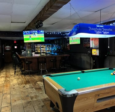 Rustic sports bar interior with a pool table, wooden floors, bar stools, and TVs showing baseball.