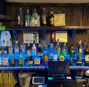 Rustic wooden bar shelves stocked with liquor bottles like Jameson and Bacardi under blue LED lights.
