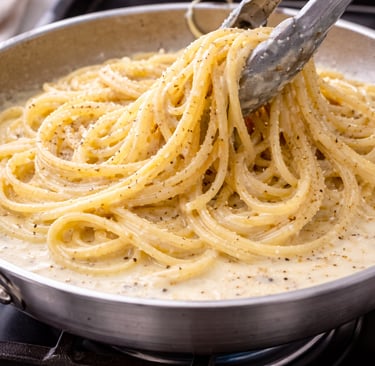 Tongs lifting creamy spaghetti cacio e pepe with cracked black pepper from a stainless steel skillet.
