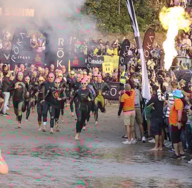 Triathletes in wetsuits start Ironman Wales swim on a beach with fire effects and a cheering crowd.