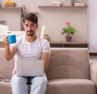 A man in pajamas sitting on a couch working on a laptop while holding a blue coffee mug.