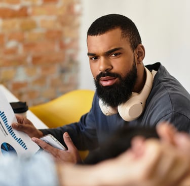 A male data analyst with a beard and headphones reviewing financial charts and business reports in a modern office.