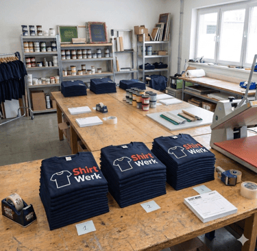 Stacks of printed navy blue t-shirts on a wooden table in a professional screen printing workshop.