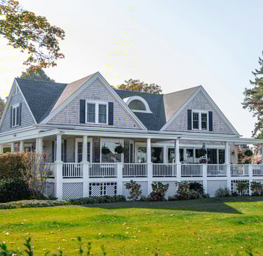A luxury Cape Cod style shingle house featuring a large wrap-around porch and green lawn.