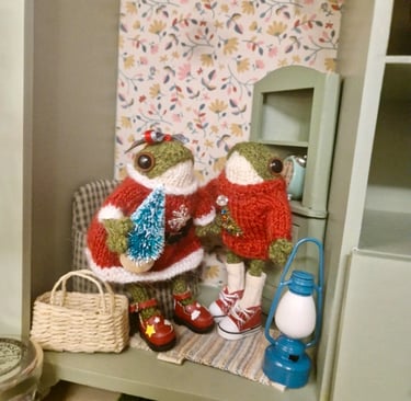 a couple of little knitted frogs wearing Christmas outfits in a doll house