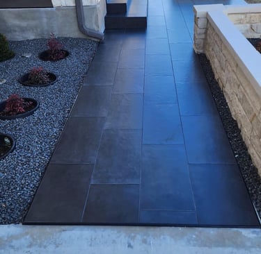 A modern dark grey porcelain tile walkway leading to a stone house entrance with xeriscape landscaping.