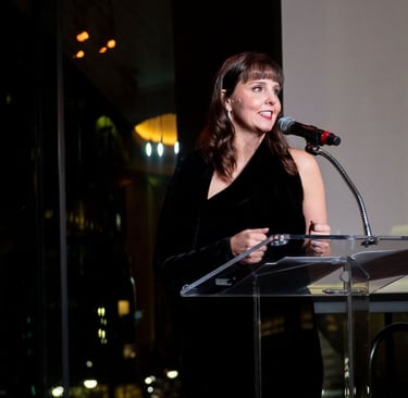 Woman speaking at a podium with a microphone at an indoor evening event, city lights visible through windows behind her.