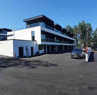 Modern white apartment building with glass balconies and paved parking area under a clear blue sky.