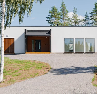 Modern white single-story house with minimalist design and gravel driveway surrounded by pine trees.