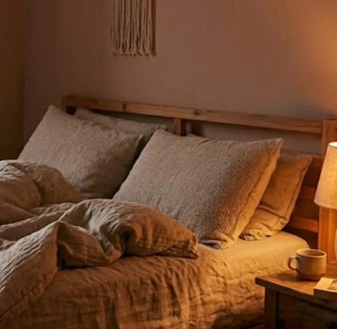 Un letto in legno con biancheria in lino grezzo, comodino in legno con sopra un libro, una tazza e una lampada con luce calda